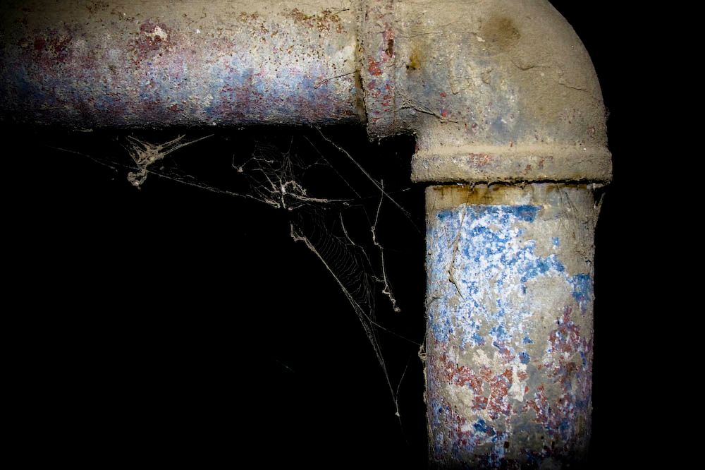 A weathered metal pipe joint with a spider's web caught in the corner, set against a dark background.
