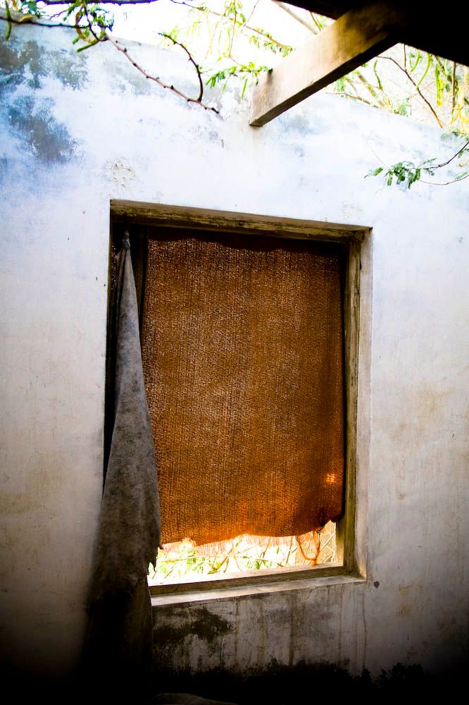 A weathered window with an orange curtain, seen from inside an abandoned building, tree branches visible through the opening.