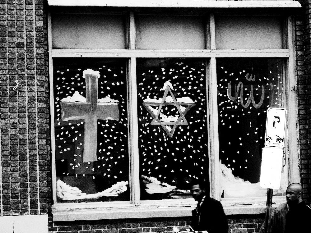 A shop window hand-painted with a cross, a Star of David, and the word Allah in Arabic script, snow falling around them, two passers-by below.