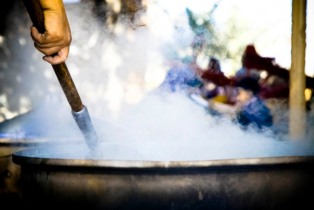 A hand stirs a large steaming outdoor cauldron with a wooden paddle, steam rising into dappled light.