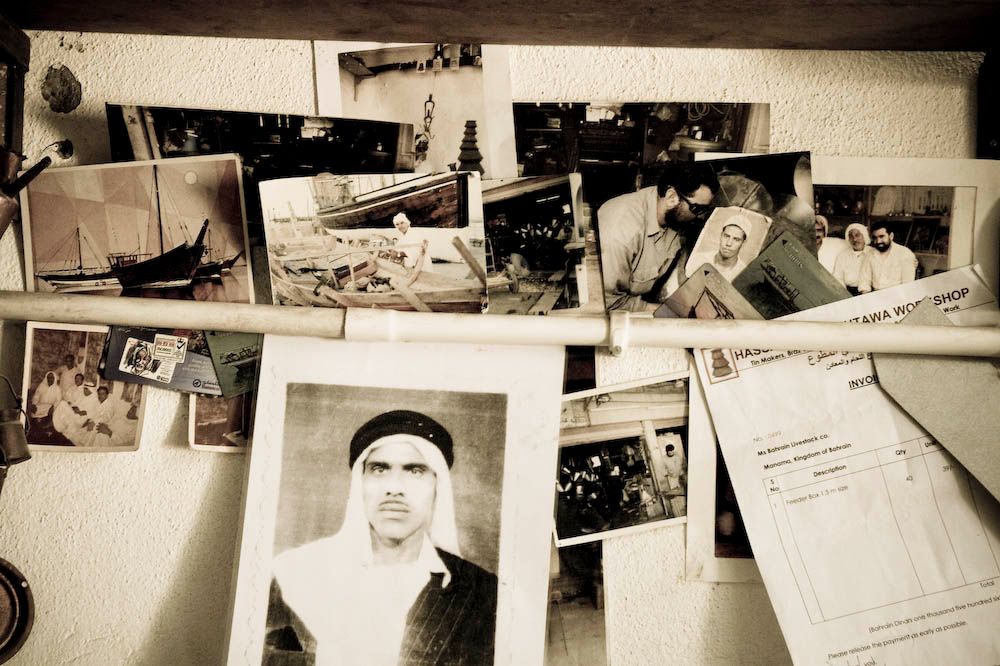 A cluttered wall of old family photographs and documents held under a pole, including boats, a workshop, and a portrait of a man in traditional dress.