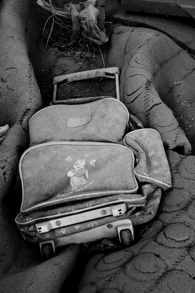 A black and white photograph of an abandoned child's Winnie the Pooh school bag, half-buried in sand and debris.