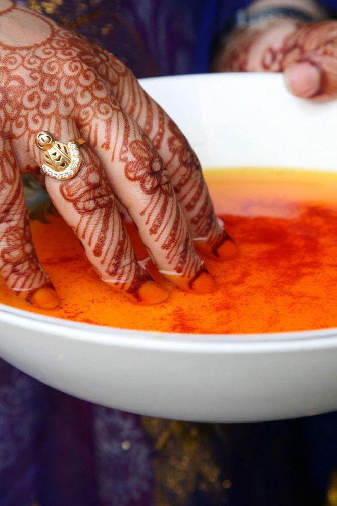 A henna-decorated hand dipping fingers into a bowl of bright orange saffron water, a gold ring visible.