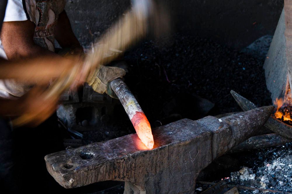 A blacksmith's gloved hand hammers a glowing red-hot piece of metal on an anvil, sparks and coal fire visible.