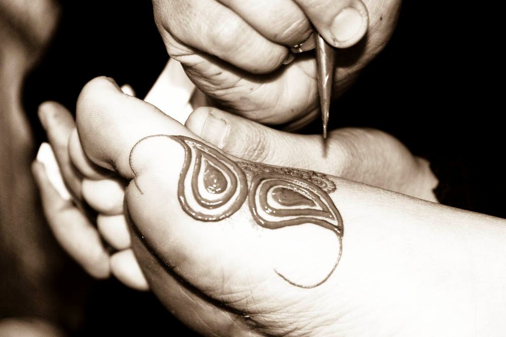 A close, sepia-toned photograph of hands applying henna in a paisley pattern to a child's foot, using a fine cone applicator.